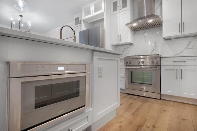 kitchen featuring wall chimney exhaust hood, light wood-type flooring, white cabinets, stainless steel appliances, and backsplash