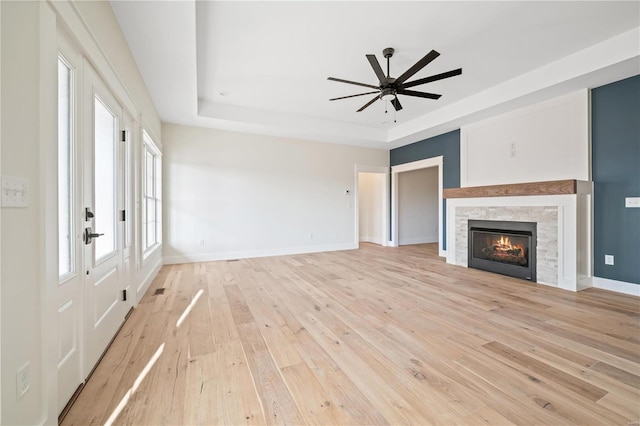 unfurnished living room featuring a tray ceiling, a stone fireplace, ceiling fan, and light wood-type flooring