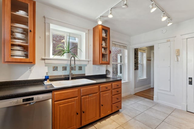 kitchen featuring brown cabinets, dark countertops, stainless steel dishwasher, glass insert cabinets, and a sink