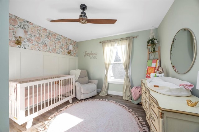 bedroom with wallpapered walls, wainscoting, ceiling fan, wood finished floors, and a nursery area