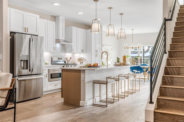 kitchen featuring white cabinets, hanging light fixtures, stainless steel appliances, a center island with sink, and wall chimney range hood