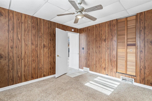 carpeted empty room with a drop ceiling, ceiling fan, and wood walls