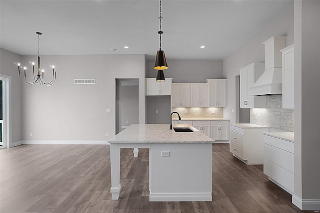 kitchen with wall chimney range hood, sink, white cabinetry, light stone countertops, and a center island with sink