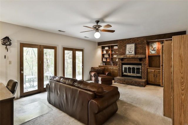 living room with a brick fireplace, built in shelves, light colored carpet, and ceiling fan