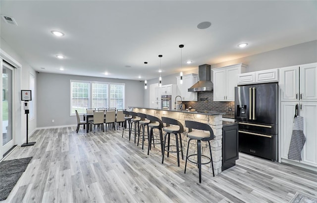 kitchen featuring wall chimney exhaust hood, high end fridge, decorative light fixtures, a center island with sink, and white cabinets