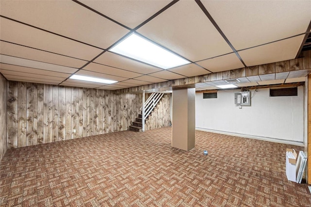 basement featuring a drop ceiling, wooden walls, and electric panel