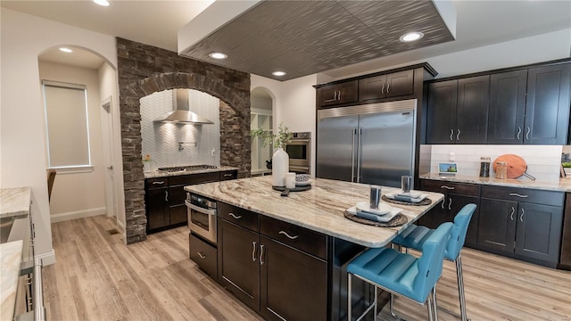 kitchen with arched walkways, wall chimney exhaust hood, appliances with stainless steel finishes, and tasteful backsplash