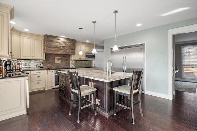 kitchen featuring sink, stainless steel appliances, an island with sink, decorative light fixtures, and dark stone counters