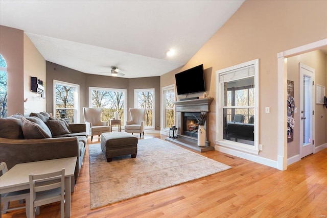 living area with a warm lit fireplace, baseboards, a ceiling fan, wood finished floors, and high vaulted ceiling