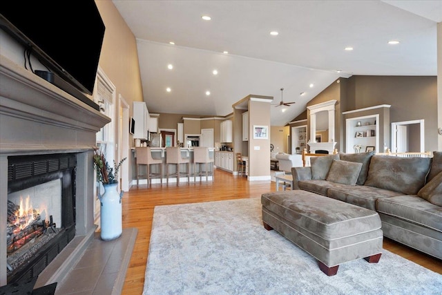 living area with a warm lit fireplace, high vaulted ceiling, light wood finished floors, and recessed lighting