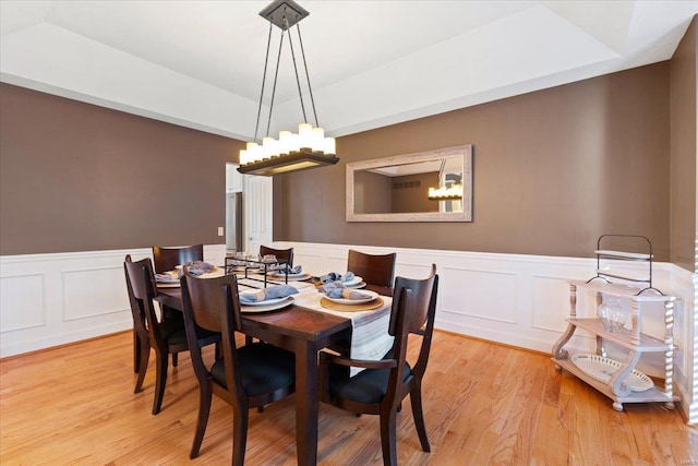 dining space with a wainscoted wall, a raised ceiling, light wood-style flooring, and a notable chandelier