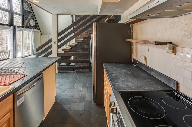 kitchen with under cabinet range hood, stainless steel appliances, backsplash, dark countertops, and stone tile flooring