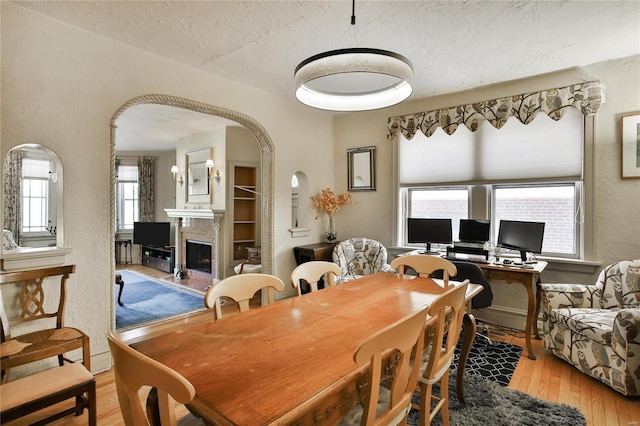 dining room featuring arched walkways, a fireplace with raised hearth, a textured wall, a textured ceiling, and hardwood / wood-style flooring