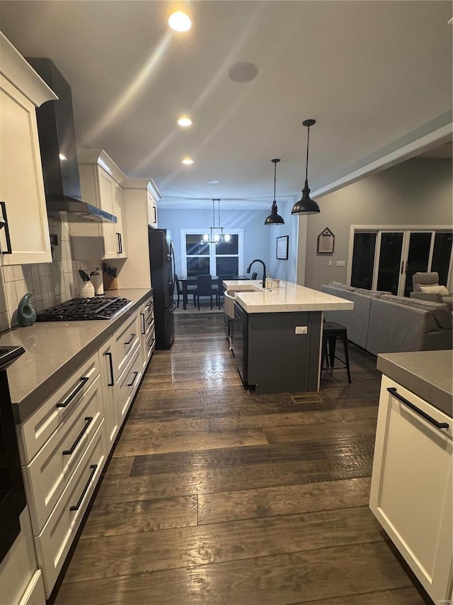 kitchen featuring black appliances, decorative light fixtures, a center island with sink, wall chimney range hood, and white cabinets