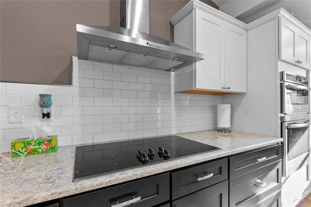 kitchen with tasteful backsplash, white cabinets, stainless steel double oven, wall chimney range hood, and black electric cooktop