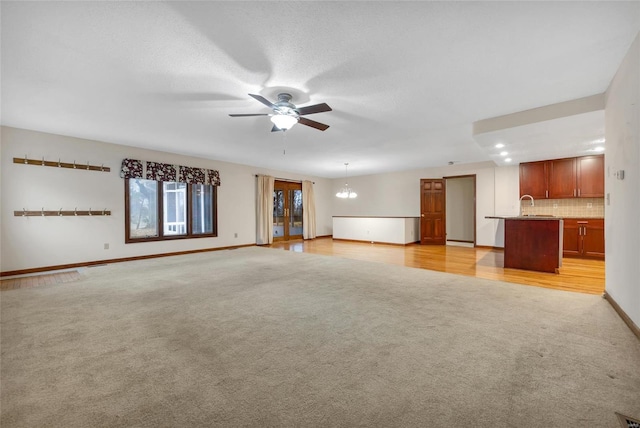 unfurnished living room with light carpet, a sink, baseboards, and ceiling fan with notable chandelier