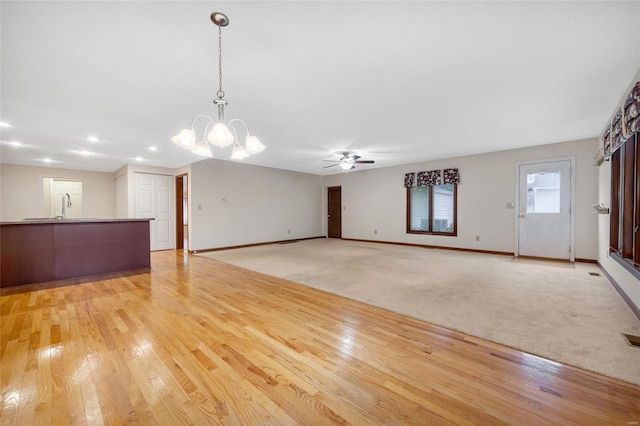 unfurnished living room with light wood finished floors, ceiling fan with notable chandelier, baseboards, and light colored carpet