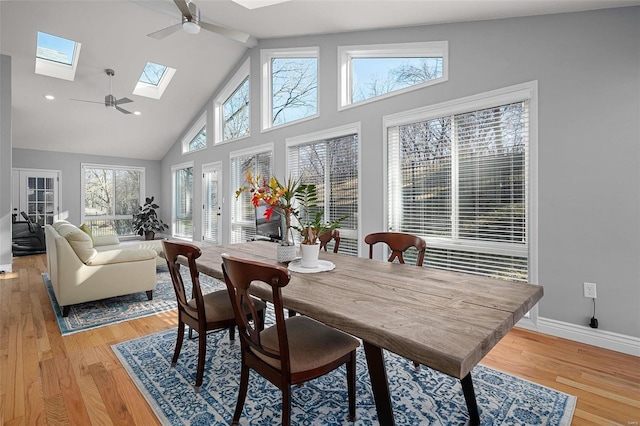 dining space with a ceiling fan, baseboards, high vaulted ceiling, light wood-style flooring, and a skylight