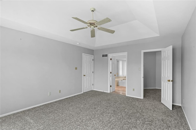 unfurnished bedroom featuring ensuite bath, a spacious closet, a raised ceiling, dark carpet, and ceiling fan