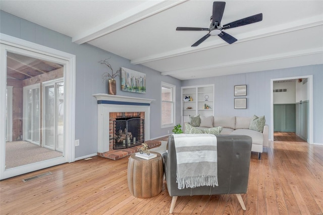 living room featuring light wood-style flooring, beamed ceiling, a brick fireplace, and visible vents