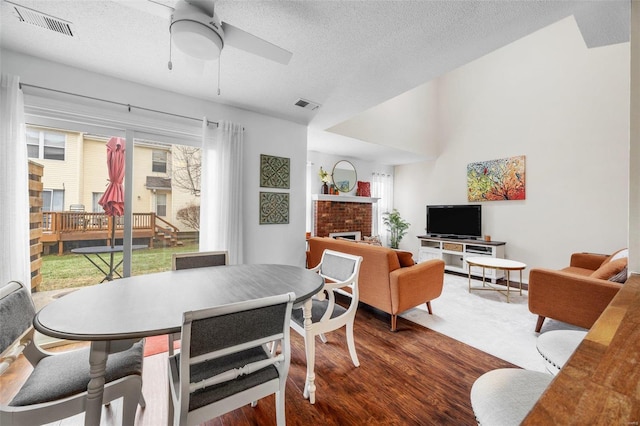 dining room with a brick fireplace, wood-type flooring, a textured ceiling, and ceiling fan