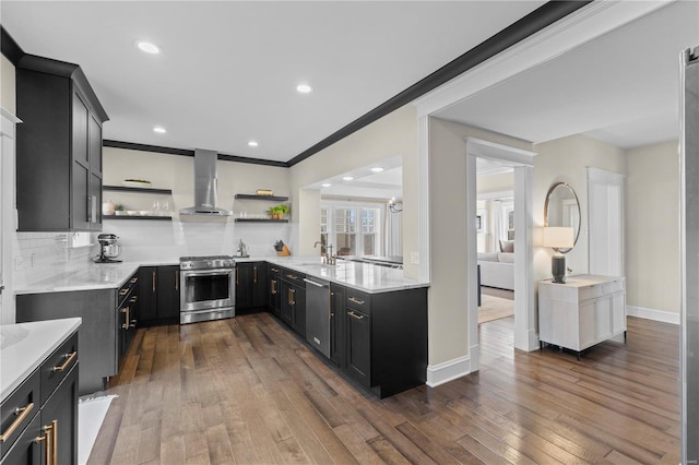 kitchen featuring open shelves, light countertops, appliances with stainless steel finishes, dark cabinetry, and wall chimney exhaust hood