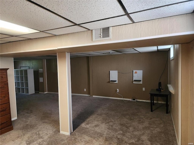 basement with a paneled ceiling, wood walls, and dark colored carpet