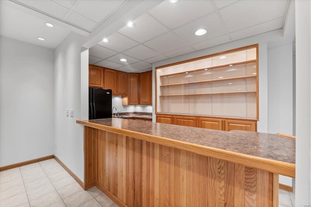 kitchen with sink, light tile patterned floors, kitchen peninsula, black fridge, and a drop ceiling