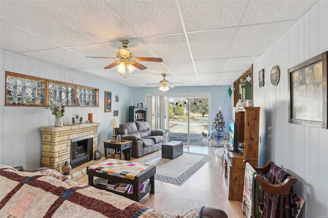 living room with a drop ceiling, a stone fireplace, light hardwood / wood-style floors, and ceiling fan