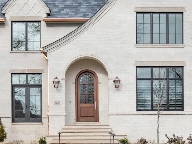 view of exterior entry with a shingled roof, french doors, and brick siding