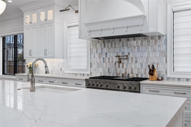 kitchen featuring light stone counters, premium range hood, a sink, white cabinetry, and glass insert cabinets