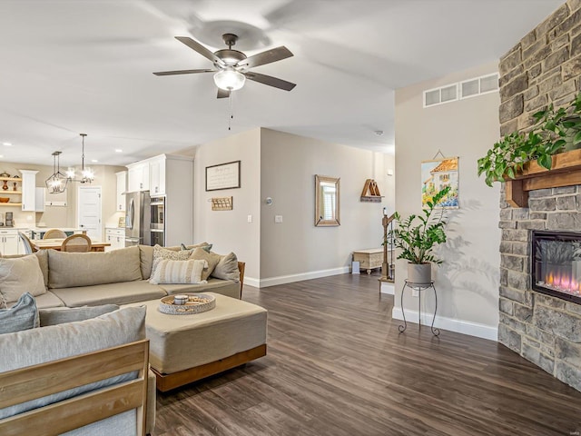 living area featuring a stone fireplace, ceiling fan with notable chandelier, dark wood-type flooring, visible vents, and baseboards