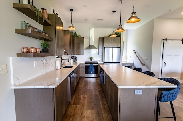 kitchen featuring wall chimney range hood, stainless steel appliances, a barn door, and light stone countertops