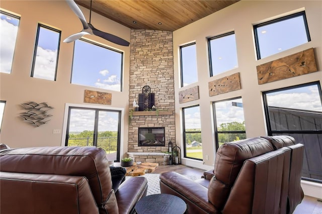 living room featuring light hardwood / wood-style flooring, ceiling fan, a towering ceiling, a stone fireplace, and wooden ceiling