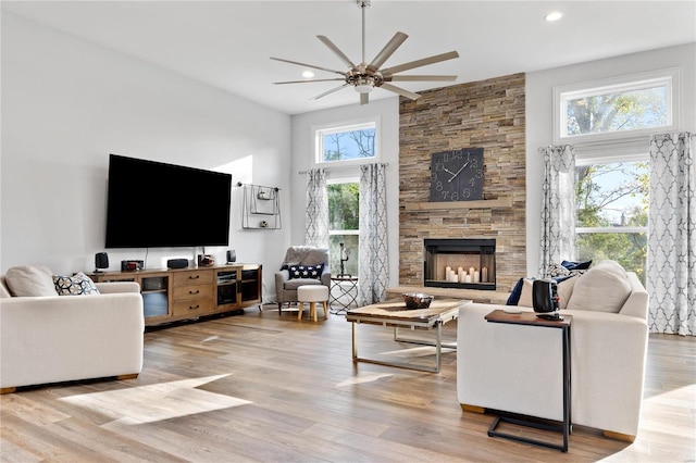 living room featuring ceiling fan, a stone fireplace, a towering ceiling, and light wood-type flooring