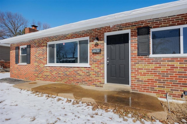 snow covered property entrance featuring a chimney and brick siding