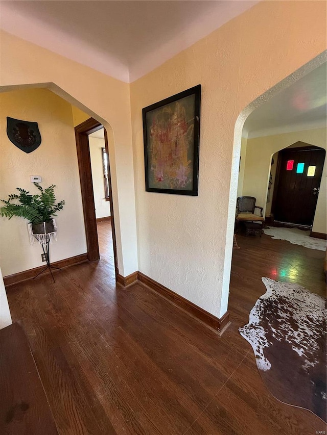 hallway with wood finished floors, arched walkways, a textured wall, and baseboards