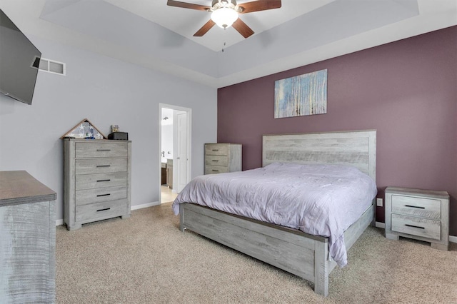 bedroom with a raised ceiling, ensuite bathroom, light colored carpet, and ceiling fan