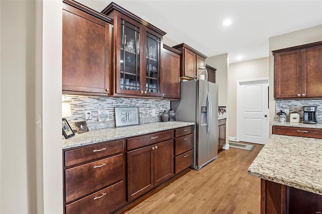 kitchen featuring light wood-style flooring, decorative backsplash, light stone counters, stainless steel fridge, and glass insert cabinets