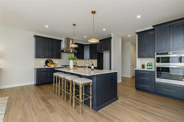 kitchen featuring a center island with sink, wall chimney exhaust hood, appliances with stainless steel finishes, a breakfast bar, and decorative light fixtures