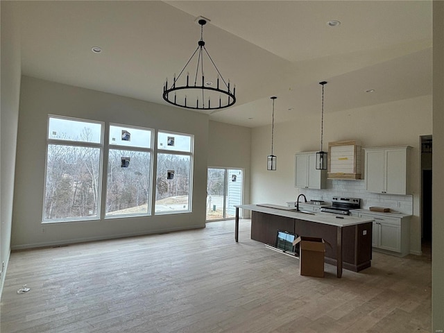 kitchen featuring a kitchen island with sink, hanging light fixtures, stainless steel range with electric stovetop, white cabinets, and light wood-type flooring