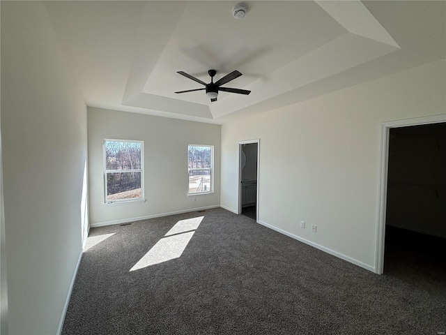 unfurnished bedroom featuring a spacious closet, baseboards, a raised ceiling, a ceiling fan, and dark colored carpet