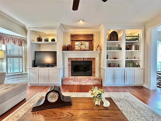 living area with wood finished floors, a ceiling fan, built in features, a brick fireplace, and crown molding