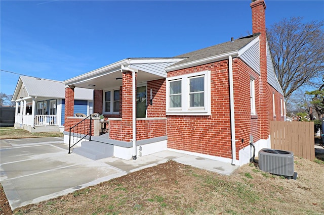 view of front of house featuring brick siding, fence, covered porch, central AC unit, and a chimney