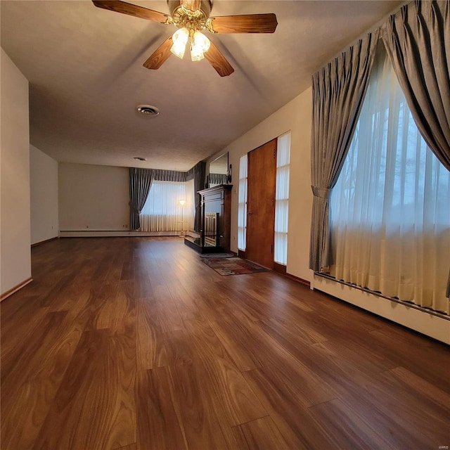 unfurnished living room featuring visible vents, a fireplace with raised hearth, a ceiling fan, a baseboard heating unit, and dark wood finished floors