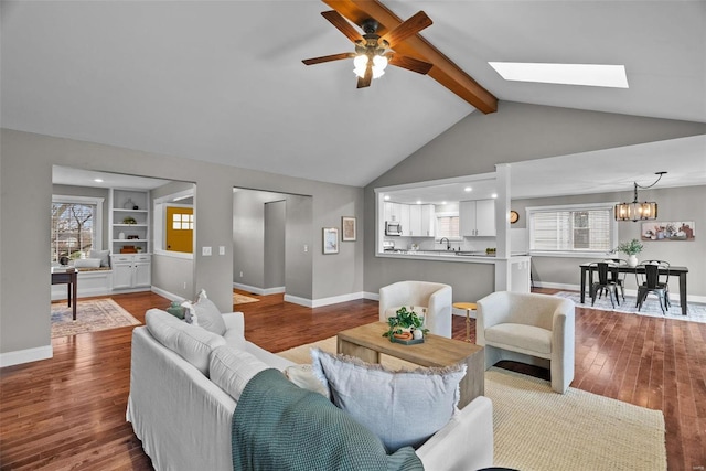 living area with built in features, wood finished floors, baseboards, lofted ceiling with skylight, and ceiling fan with notable chandelier