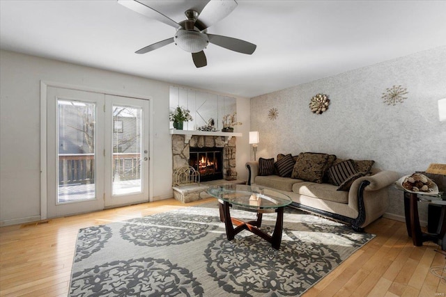 living room featuring wallpapered walls, baseboards, ceiling fan, a stone fireplace, and light wood-type flooring