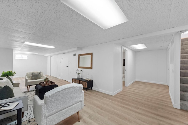 living area featuring stairs, light wood-style floors, baseboards, and a drop ceiling