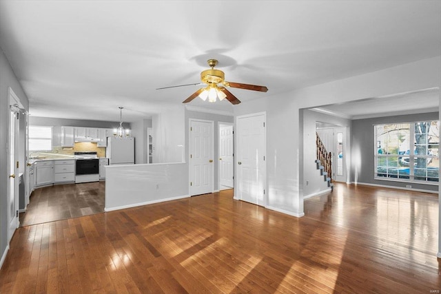 unfurnished living room featuring stairway, baseboards, dark wood finished floors, and ceiling fan with notable chandelier