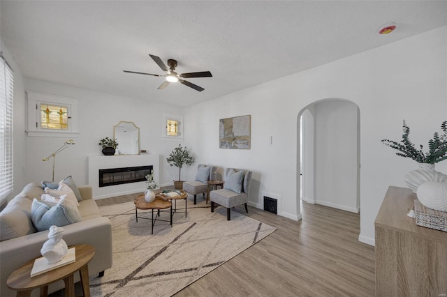 living room featuring arched walkways, a ceiling fan, a glass covered fireplace, wood finished floors, and baseboards